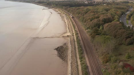 Stationary Aerial shot of a train traveling along railway tracks with a shoreline and Motorway on either side.