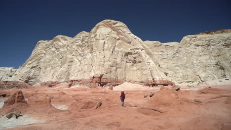 lonely woman with backpack walking in dry desert landscape under sandstone hills on hot sunny day, back view