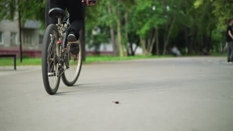 back view of cyclist wearing black sneakers and trousers, pedaling bicycle steadily along paved path, with blurred background of another individual standing near bicycle surrounded by greenery