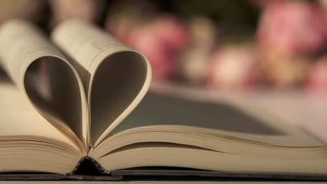 open book with pages forming heart shape and rose flowers bouquet on a table