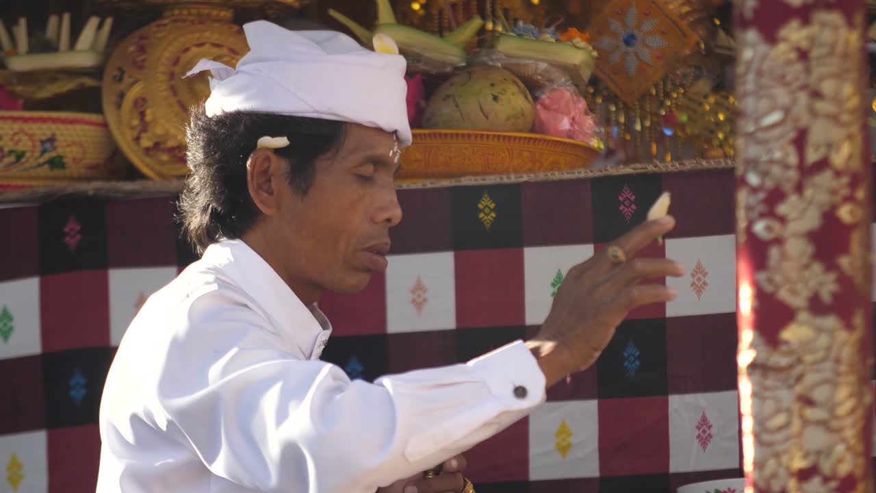 Hindu priest sitting at altar performing a religious ritual using flowers, rice and water. Smoke from burning incense and man praying. Hinduism and cultural practices of Indonesia.