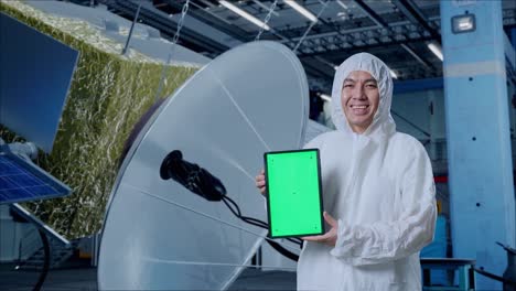 científico masculino asiático sonriendo y mostrando la tableta de pantalla verde mientras está de pie con la construcción de la nave espacial