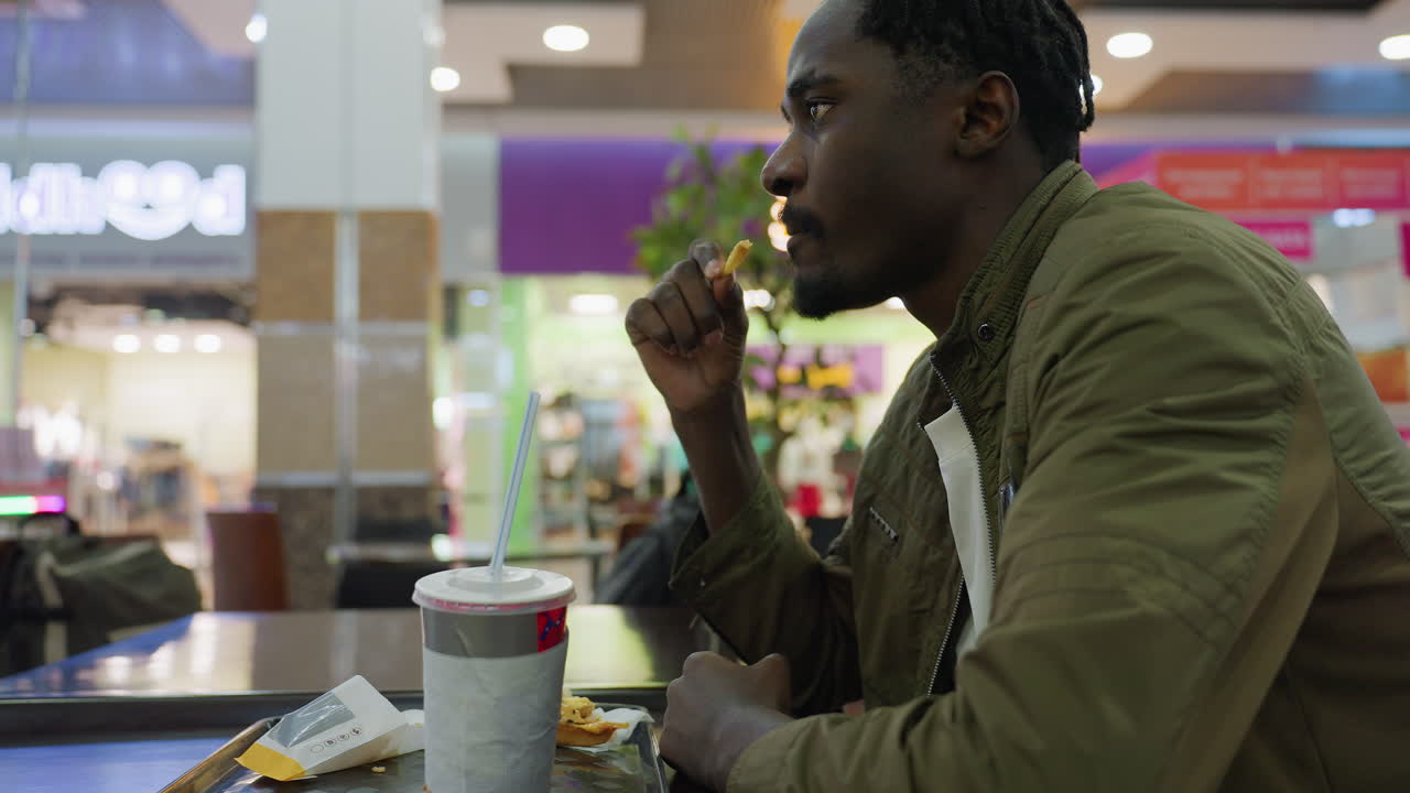 Side view of young man in olive jacket sitting alone at food court table eating fries, with paper cup and empty food pack on tray in front of him surrounded by blurry mall background