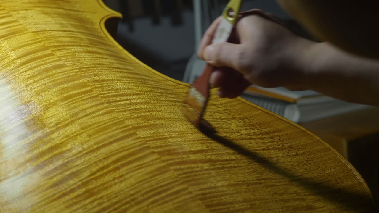 Craftsman’s hand uses paintbrush spreading linseed oil, mastic, and lac natural varnish on the marbled back of a cello, revealing wooden vivid flames and unique patterns under precise lighting