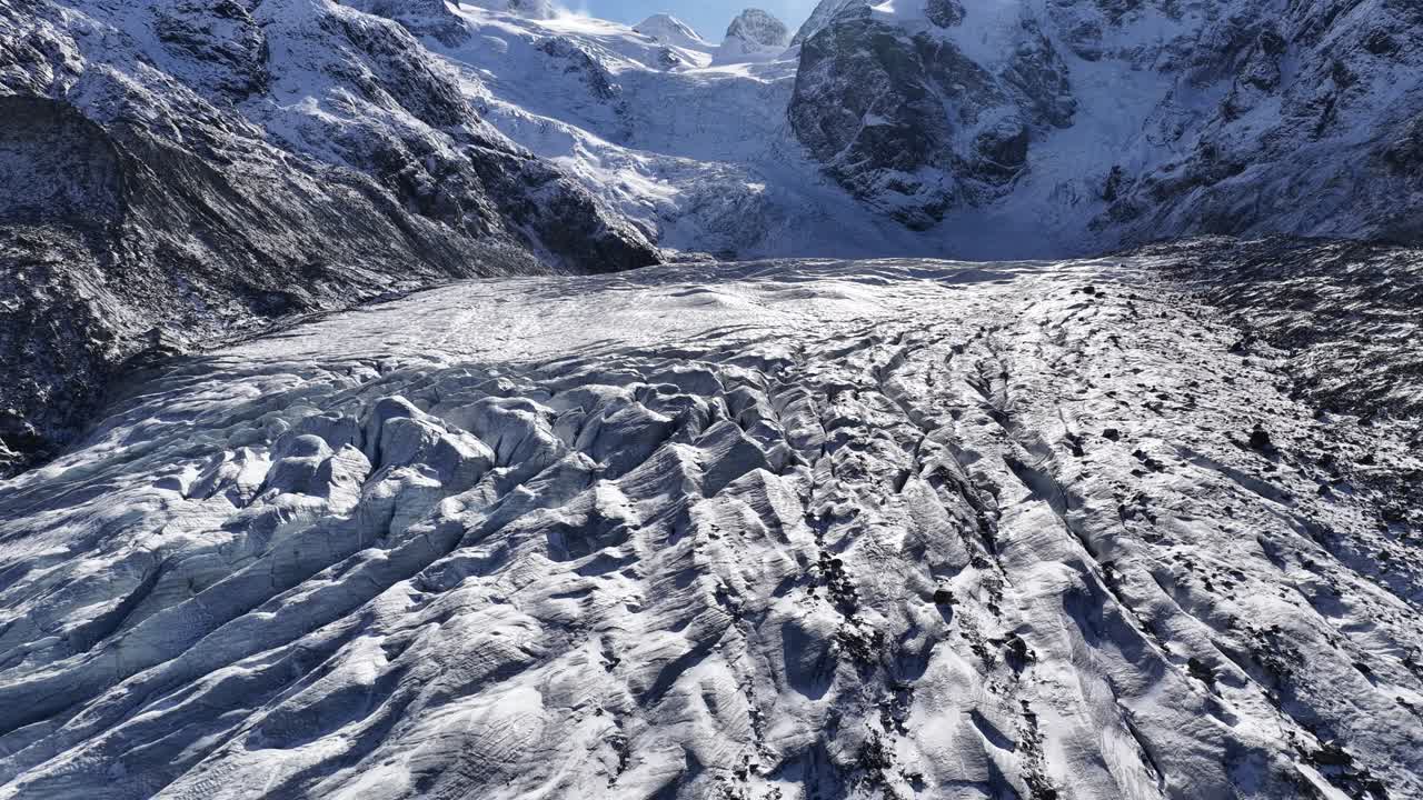 Drone flying forward over a snowy glacier valley with rugged ice patterns and surrounding mountains in Switzerland