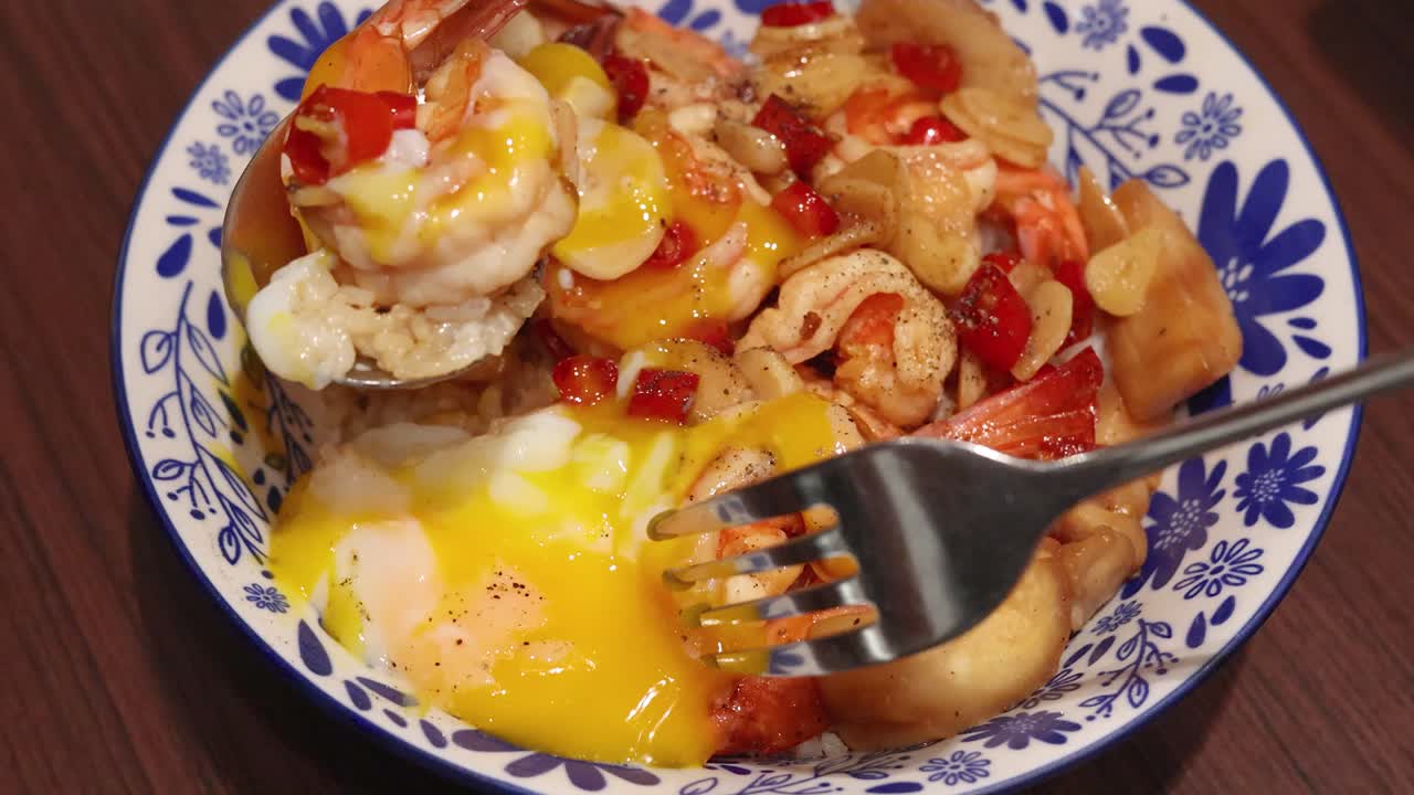 A fork pierces a soft-boiled egg atop Thai shrimp, garlic, chili, and rice, releasing yolk across a vibrant meal under warm indoor lighting