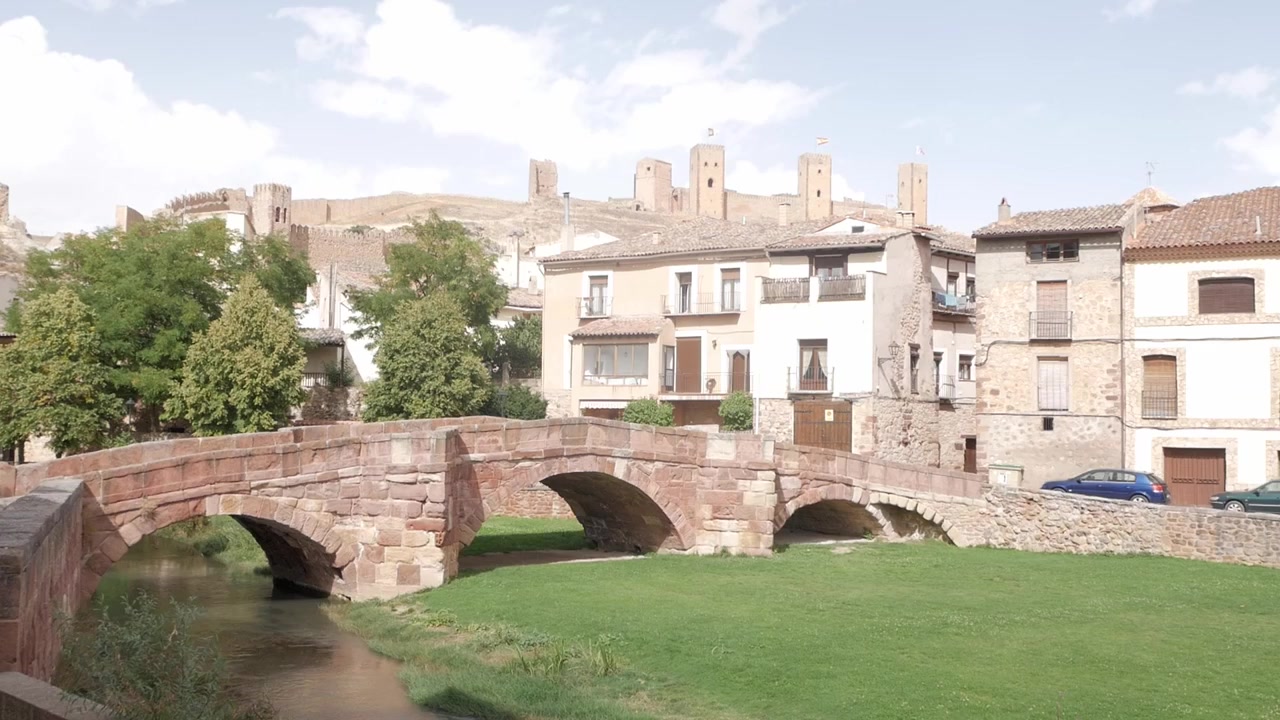 Puente de piedra histórico que cruza un pequeño río con un pueblo medieval y un castillo detrás