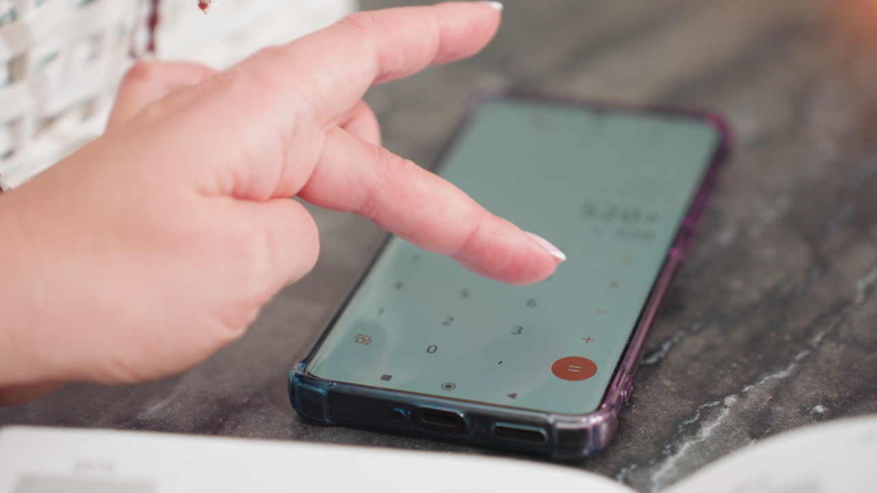 Close up of woman with polished nails using calculator app on smartphone placed on marble table while surrounded by study materials and items, finger pressing keypad with focus on solving equation