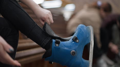 close-up of a person wearing blue ice skates while adjusting them, with a focus on the hands and black trousers, with a blur background