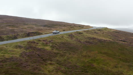 Verfolgung-Eines-Autos,-Das-Auf-Einer-Schmalen-Straße-Auf-Dem-Land-Gegen-Ein-Anderes-Auto-Fährt.-Wiesen-Und-Wiesen-Unter-Bedecktem-Himmel.-Irland