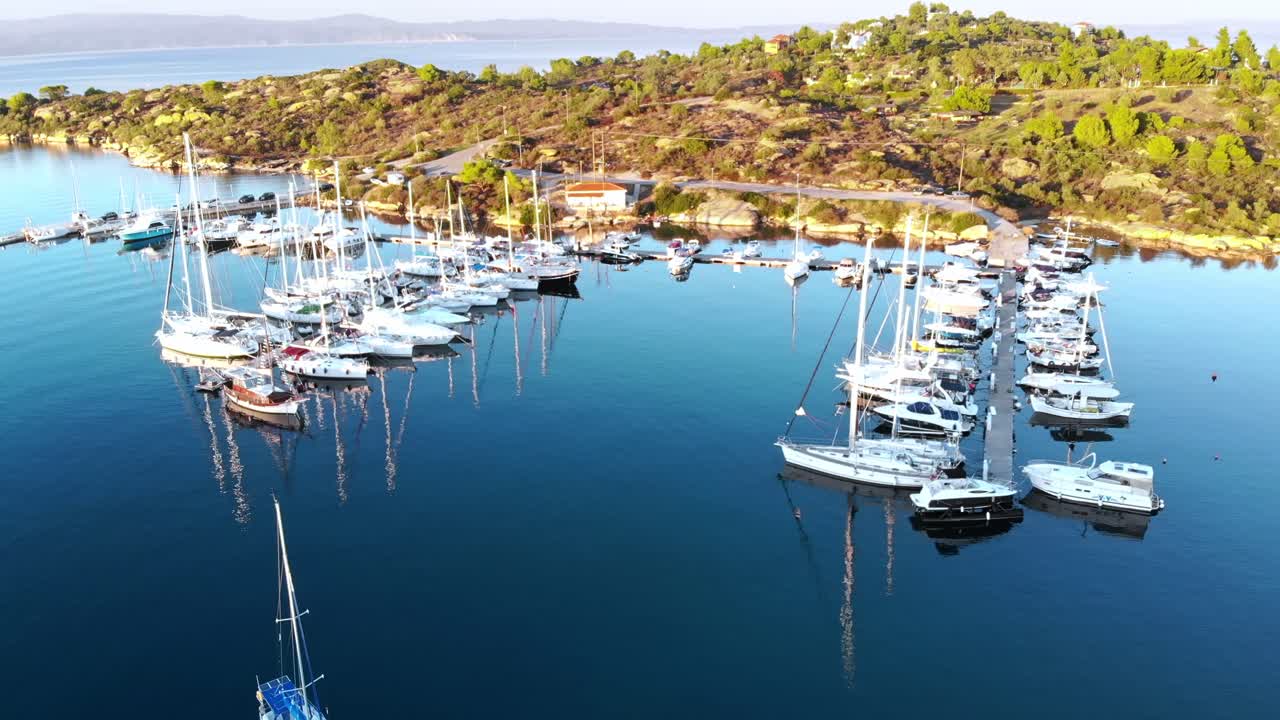 Aerial drone view of Aegean sea port with multiple moored yachts at piers, greenery, blue water, sunset, Greece