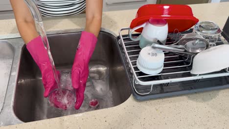 person wearing pink rubber gloves washes and rinses glassware at a stainless steel kitchen sink under bright lighting, with clean dishes drying nearby