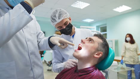 male patient sitting in dentist chair with his mouth open. two students bent over him, point at something in his mouth and comment. classroom background.