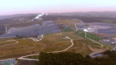 Aerial-Dolly-Over-Waste-Recycling-Landfill-With-Smoke-Rising-From-Building-In-Distance