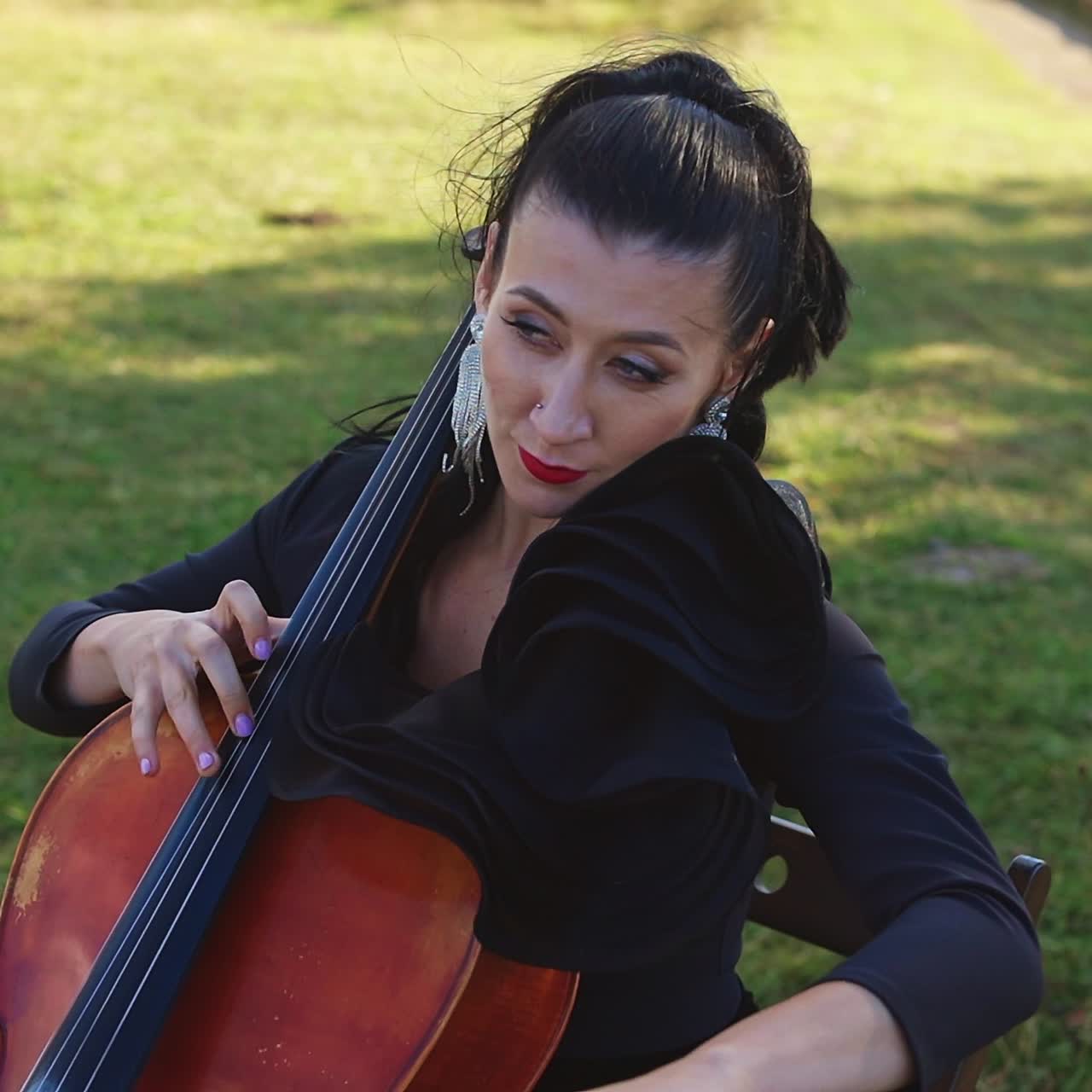 Lady wearing black dress, long earrings and red lipstick playing cello outdoors. Female musician performing music and enjoying the process. Nature backdrop