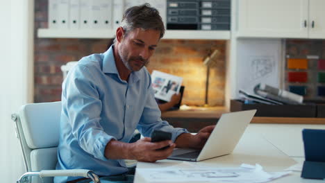 Reifer-Lächelnder-Geschäftsmann-Mit-Laptop,-Der-Am-Schreibtisch-Im-Büro-Arbeitet-Und-Anrufe-Mit-Dem-Mobiltelefon-Entgegennimmt