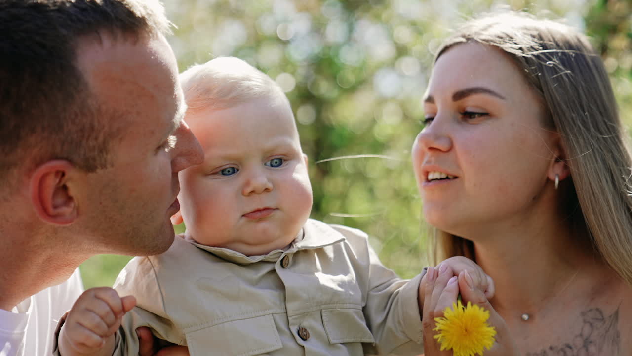 Mother, father and little baby together outdoors on sunny day. Caucasian couple kisses their beloved child on the cheeks. Kid holding a flower. Close up.