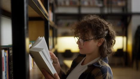 Confident-girl-with-curly-hair-with-glasses-in-a-checkered-shirt-reads-a-book-near-a-shelf-with-books-in-a-university-library