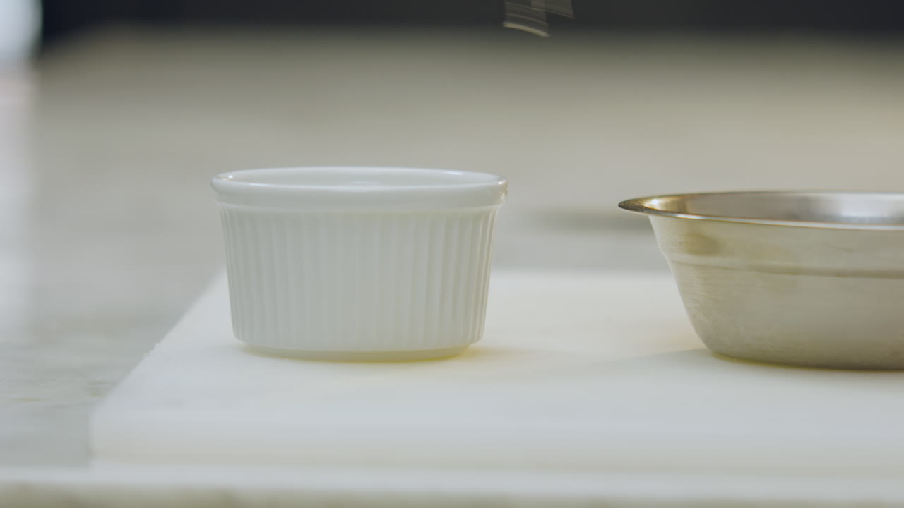 Close-up of a chef's hand using chef's tongs to place apple slices in a white ceramic mold, next to a metal container on a white chopping board