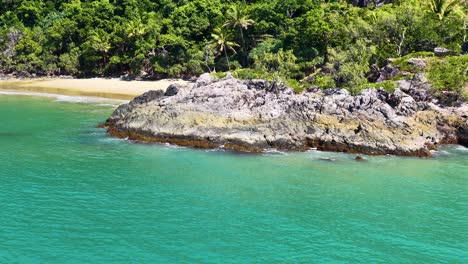 aerial footage pans over a rocky outcrop surrounded by turquoise water and lush rainforest coastline under bright daylight, highlighting natural beauty and vibrant colors