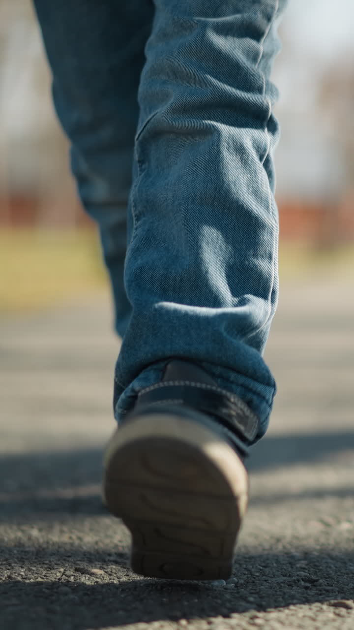 un primer plano de un niño con pantalones vaqueros y botas caminando por una acera con alguien, el niño está levantando una pierna en medio del paso