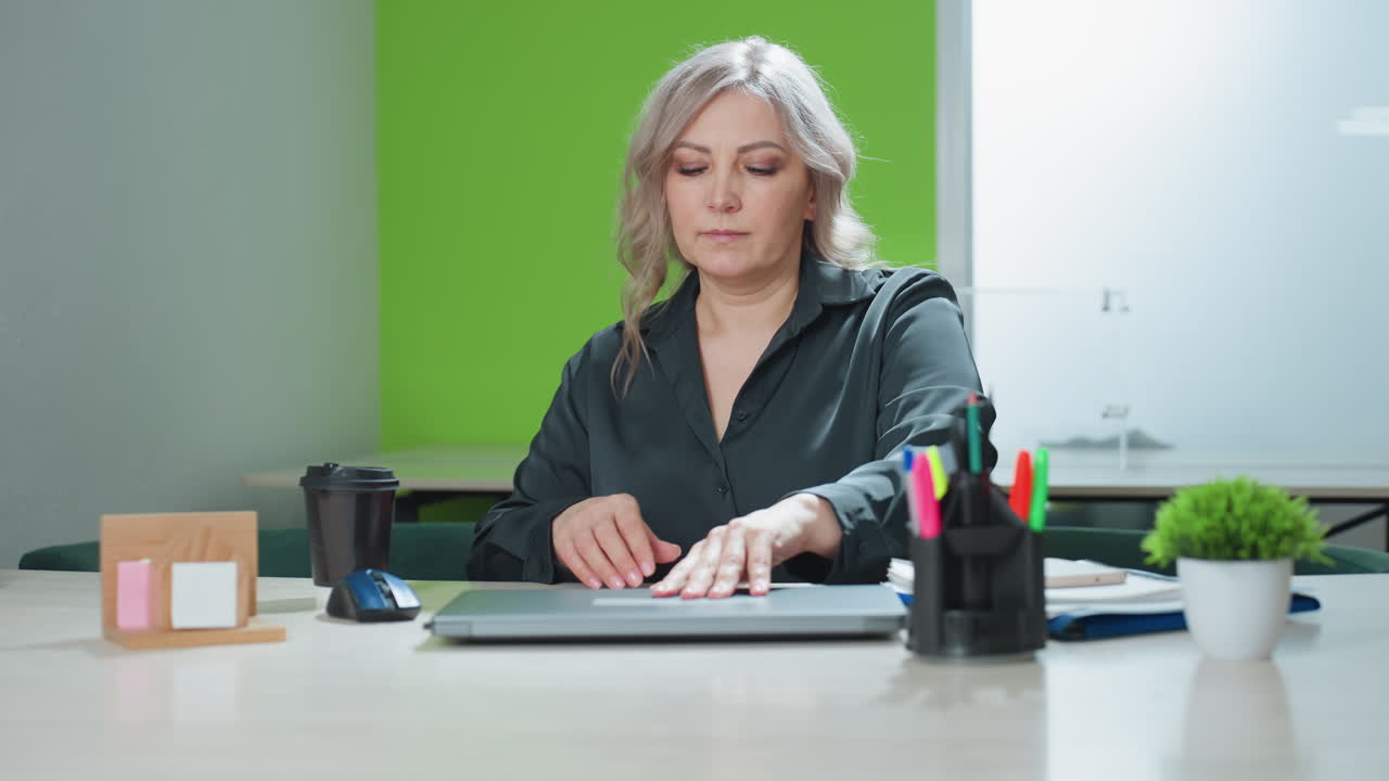 Real estate professional in dark blouse places folder on table, adjusts coffee cup, and prepares to open laptop in bright modern office setup with stationery