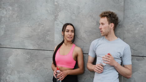 happy sportive couple leaning against a wall, holding drinking bottle and taking a break during running session in the city