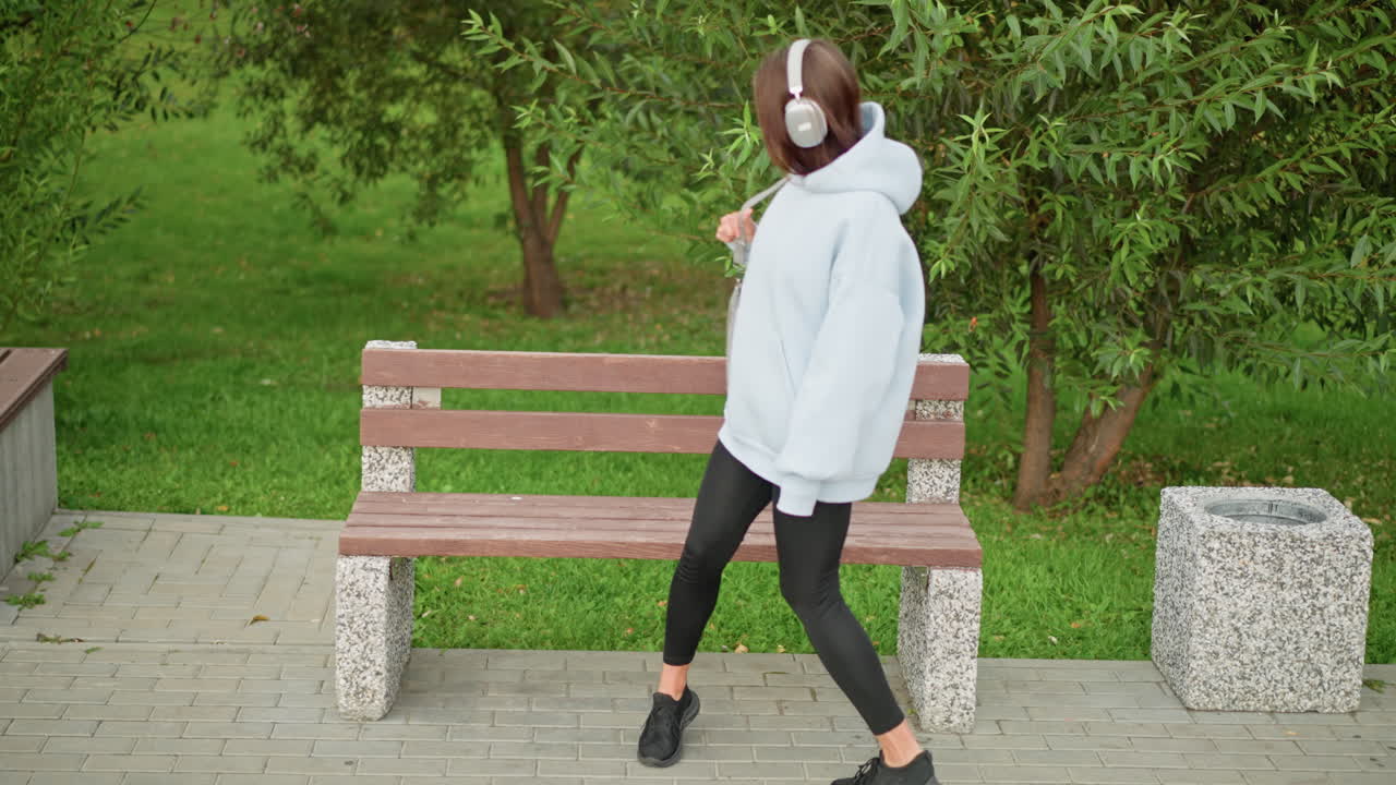 Aerial view of woman walking in park, heading to sit on concrete bench, then opening handbag, lush green leaves and calm atmosphere in peaceful park setting, ideal for relaxation and nature scenes