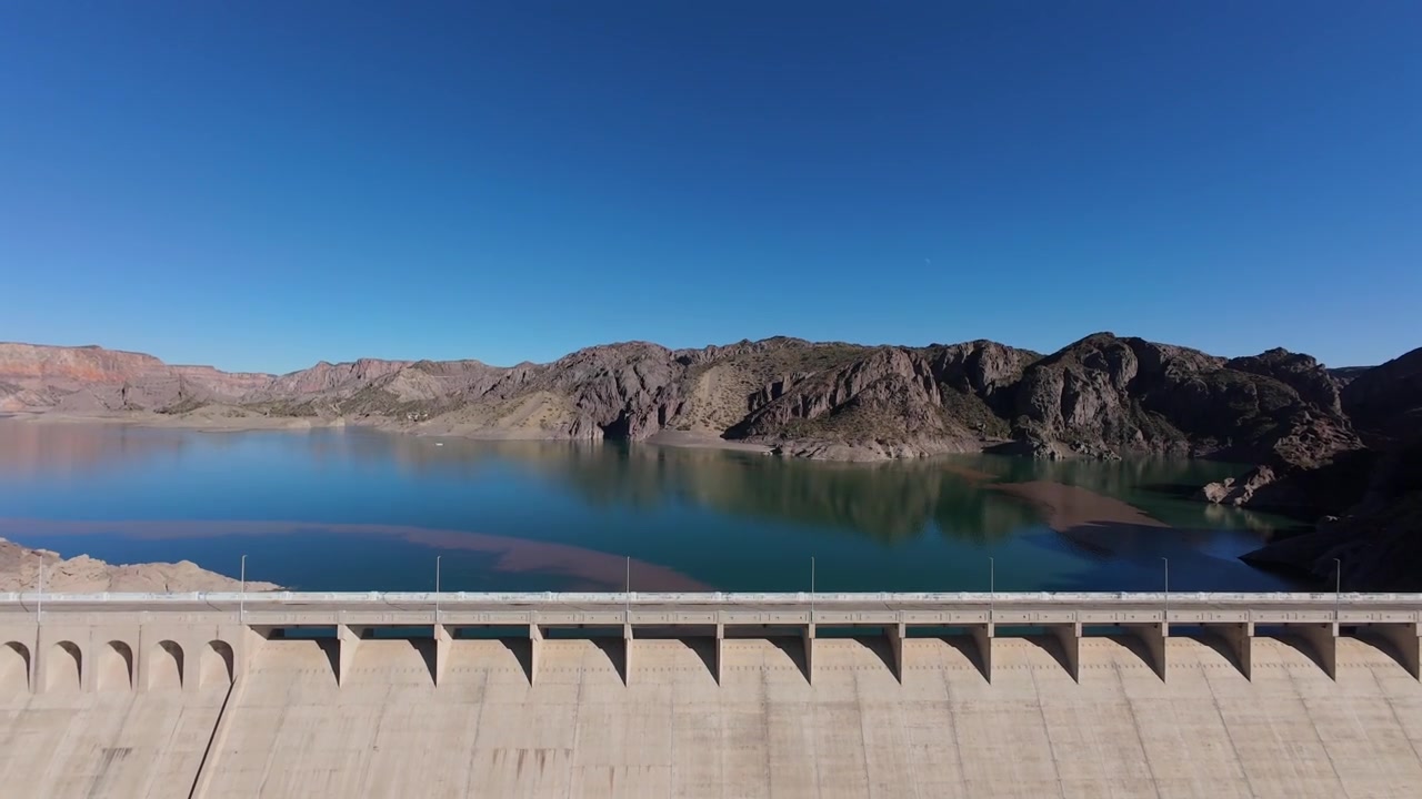 Spændende FPV-dronedyk over den massive betonmur ved Valle Grande-dæmningen i Mendoza, Argentina, der afslører Atuel-flodens fantastiske smaragdgrønne vand nedenfor.