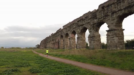 Detail-of-an-aqueduct-from-ancient-Rome-in-parco-degli-acquedotti-in-the-outskirts-of-the-capital-of-Italy-with-a-jogger-running,-pan-movement-and-wide-angle