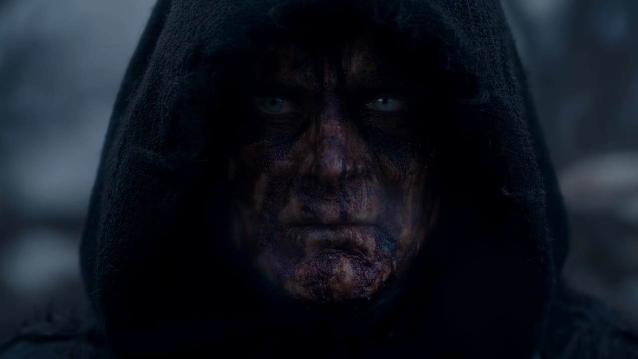 Figura incappucciata che fissa con lo sguardo, avvolta in un mantello scuro, la nebbia rivela occhi pallidi e trucco sfregiato nel bosco