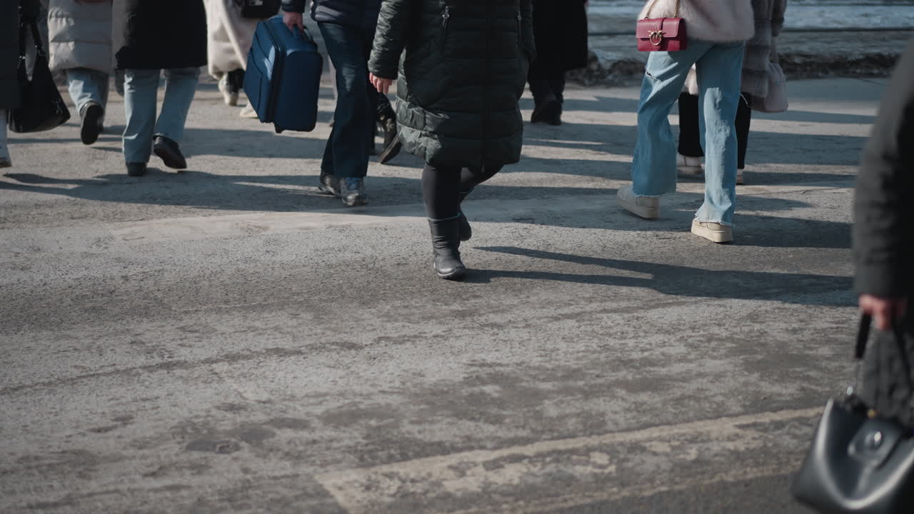 low angle crowd in winter jackets with backpacks and shopping bags walking sideways on city sidewalk, boots and shadows on asphalt, urban commute movement, cold day pedestrian traffic during morning