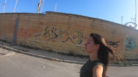 woman walking along a sunny street with graffiti and cars near a marina
