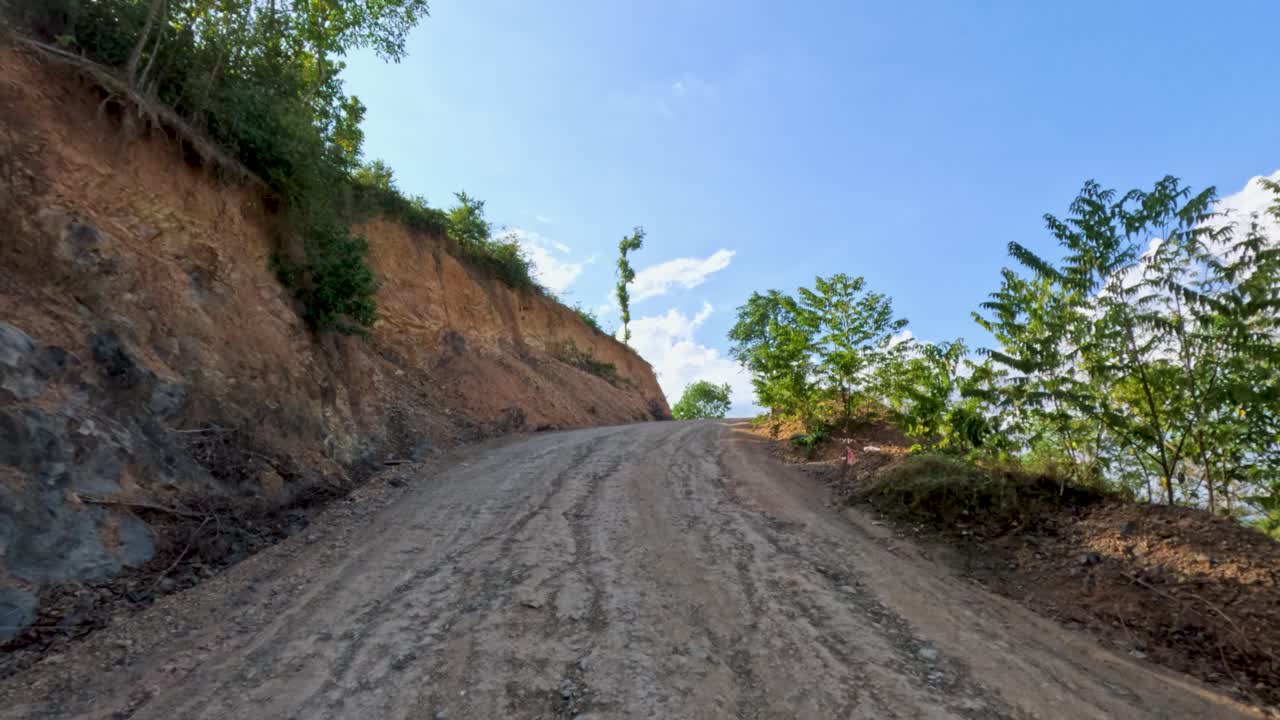 Vehicle ascends rugged dirt road, hillside cut, midday sunlight, steady forward camera, tropical environment