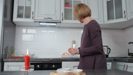 mujer elegante en vestido elegante arregla los platos en la mesa de la cocina para el almuerzo, una vela roja arde en el fondo, creando una atmósfera acogedora y cálida en un entorno doméstico moderno