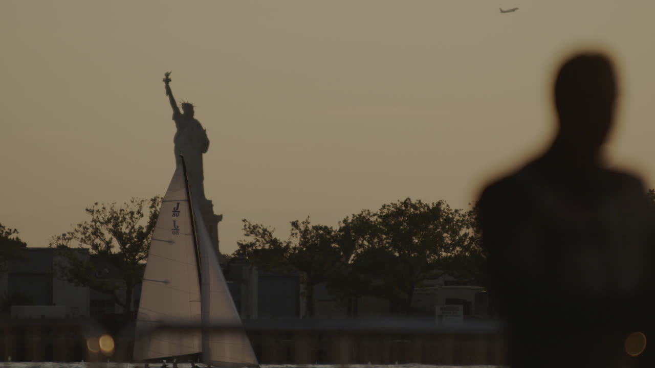 Locked off slow motion shot at dusk looking out towards the statue of liberty with sailing boat floating by and airplane taking off in the background