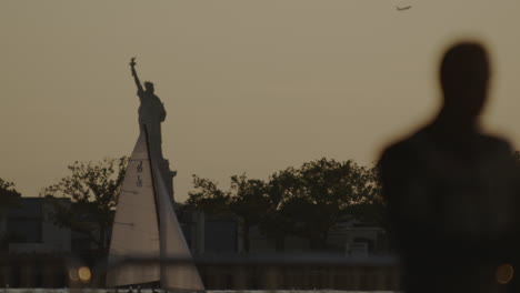 locked off slow motion shot at dusk looking out towards the statue of liberty with sailing boat floating by and airplane taking off in the background