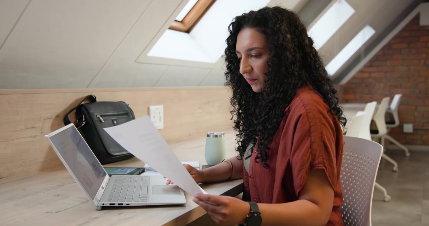 African American woman referencing laptop writing typing and comparing documents on screen at loft