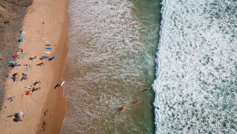 An-aerial-drone-shot-of-wave-hitting-a-beach-where-people-are-swimming-and-surfing-in-Algarve,-Portugal