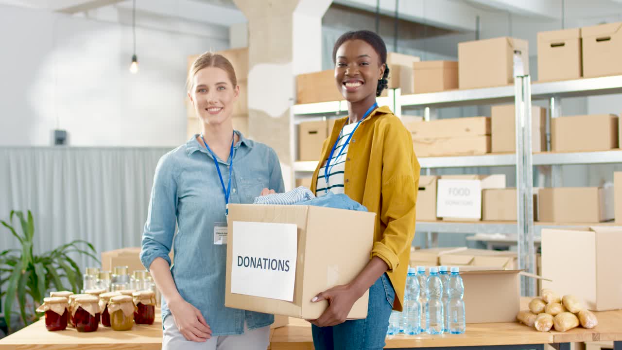 Cheerful young beautiful female volunteers holding donation box with clothes and smiling to the camera in charity warehouse