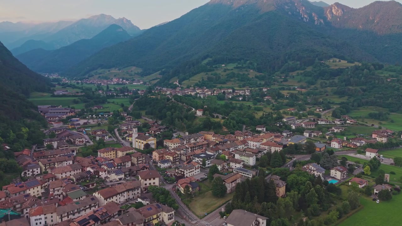 Vista aerea con drone sui tetti del villaggio di Bezzecca con chiesa gialla, valle alpina, calda luce dorata sulle cime delle montagne e cielo al tramonto dai colori pastello sullo sfondo.