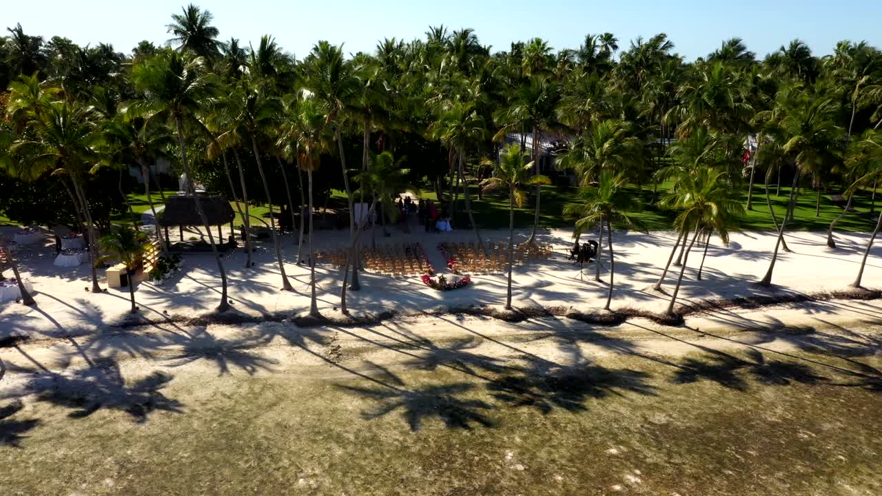 Angled aerial orbit above water to resort clearing and bordering trees with charis arranged for wedding