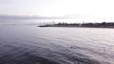 Man-Swimming-In-Ocean-Over-Melbourne-City-Drone