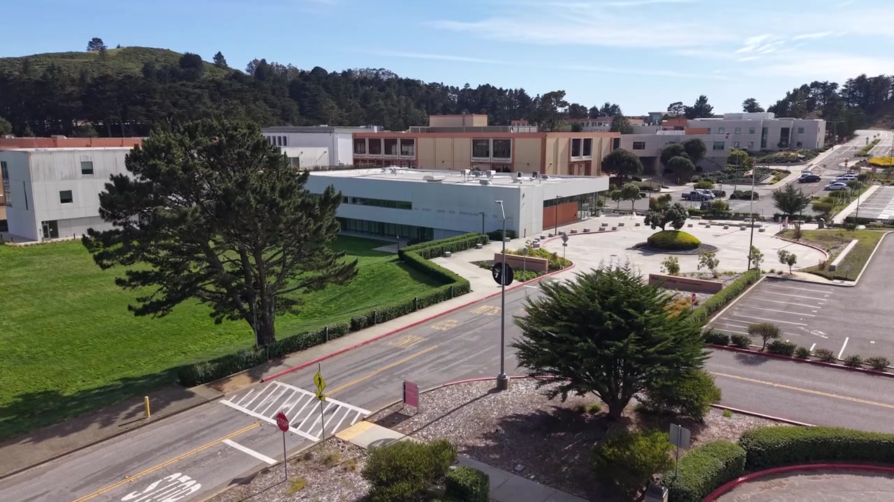 Vista de dron ascendente sobre el estacionamiento vacío y los edificios modernos del campus de Skyline College en San Bruno, California, en una tarde soleada y luminosa con cielo azul y paisaje verde