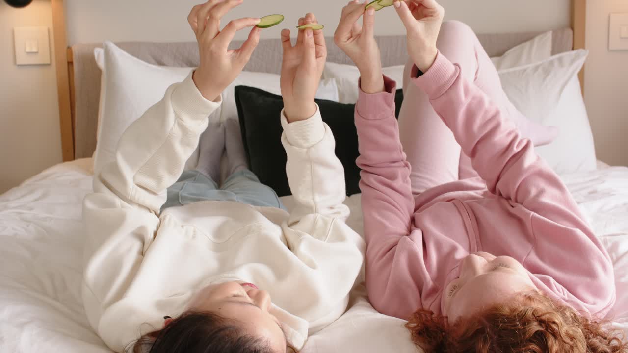 Amigas deitadas em uma cama com travesseiros, usando moletons rosa creme e segurando fatias de pepino para um tratamento de spa