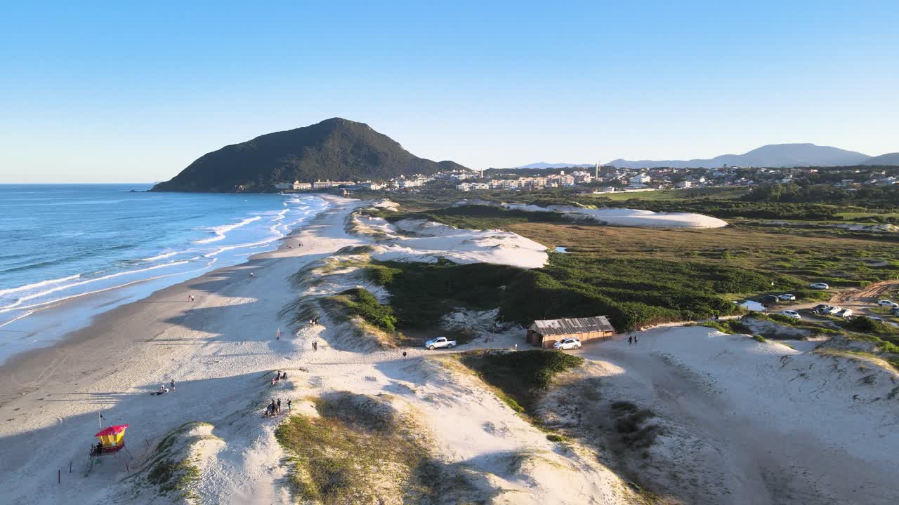 Ripresa aerea orbitale cinematografica di Praia do Santinho a Florianópolis, Brasile, che rivela dune di sabbia bianca, onde oceaniche turchesi e la montagna Morro dos Ingleses in una giornata di sole