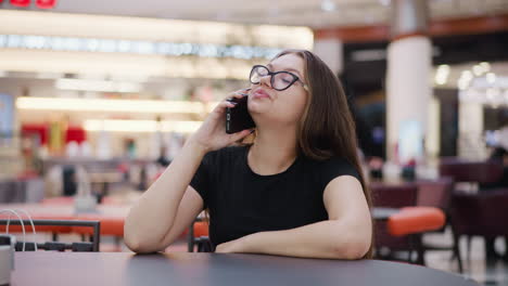 mujer joven con camiseta negra y gafas hablando por teléfono, sentada en la mesa de la corte de alimentos, sonriendo mientras mira a su alrededor con luces bokeh en el fondo