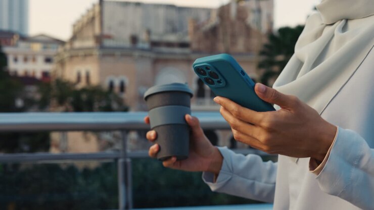 Nahaufnahme von Händen, die ein Smartphone und einen Reisebecher in einer städtischen Umgebung halten