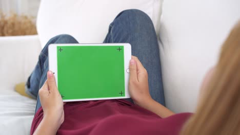 mujer joven asiática usando un dispositivo de tableta negra con pantalla verde. mujer asiática sosteniendo una tableta, desplazando páginas mientras está sentada en el sofá en la sala de estar. clave de croma.