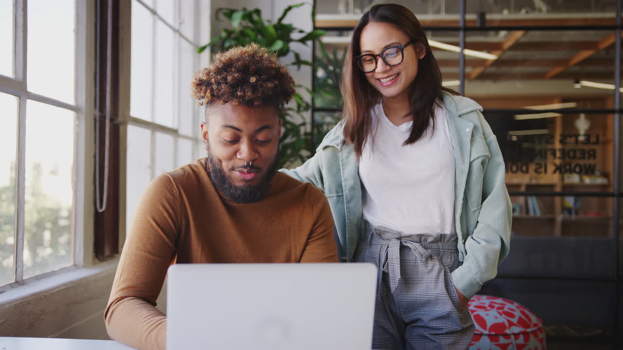 Two millennial creatives looking at a laptop together at a desk in an office, close up